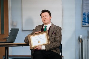 Daniel Rigby holding a certificate on stage in a scene from Accidental Death of an Anarchist