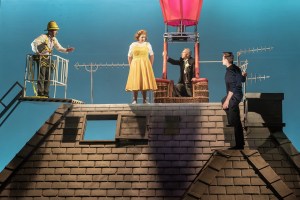 Four actors are pictured on stage on a rooftop with one inside the basket of a hot air balloon