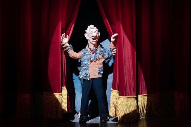 A Spitting Image puppet of Ian McKellen holds the curtain open on stage