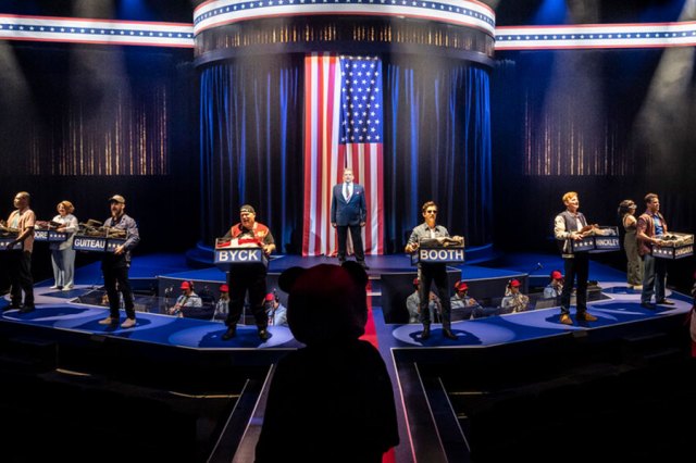 A photo showing a stage with performers standing at lecterns, in contemporary clothing. A US flag is draped behind them 