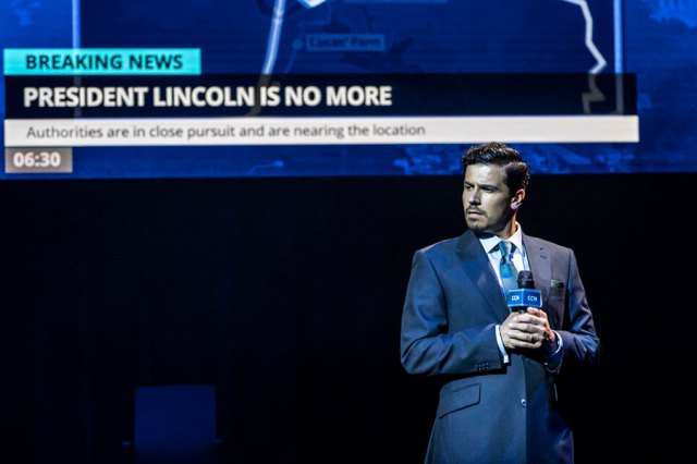 Liam Tamne stands in a suit on stage in Assassins