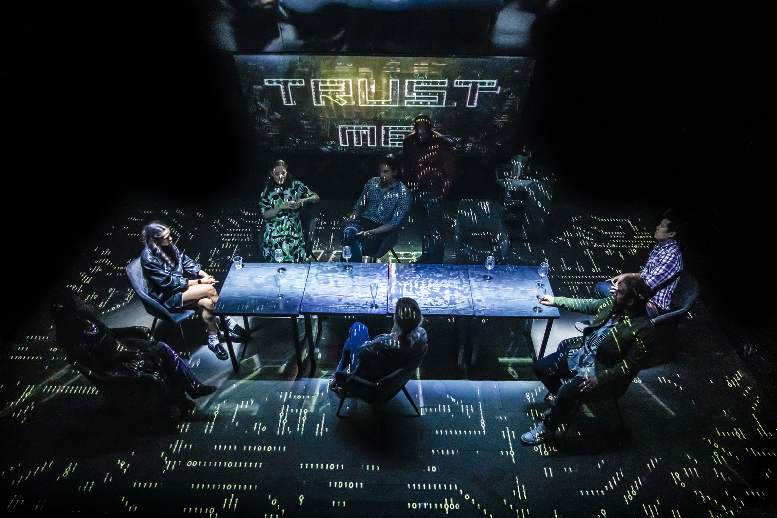 Cast members sit around a table on a stage that resembles a circuit board that displays the words "Trust Me" above them
