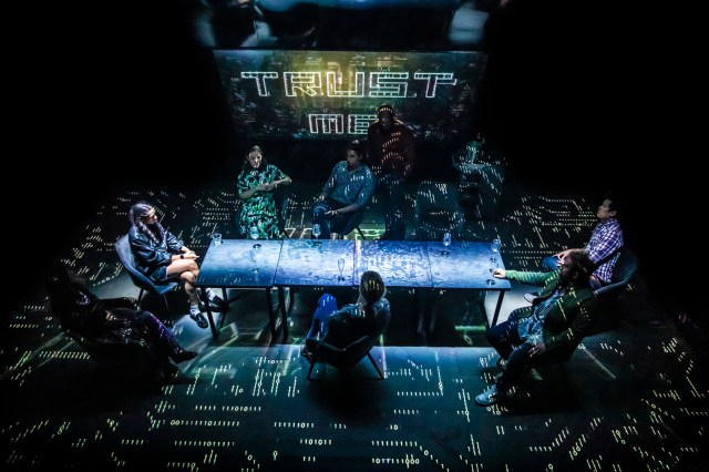 Disruption Pamela Raith Photography Cast members sit around a table on a stage that resembles a circuit board that displays the words "Trust Me" above them