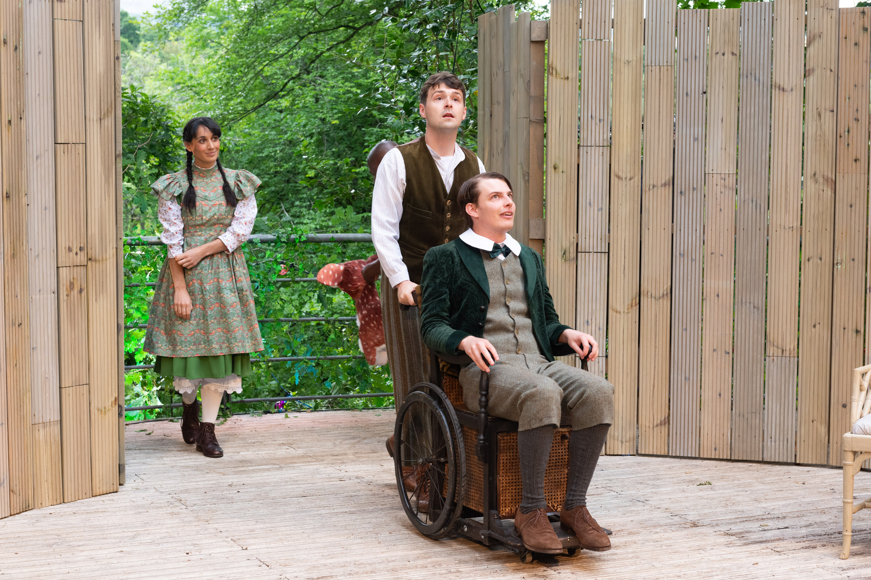Three actors in period costumes on stage, one of whom is sat in a wheelchair