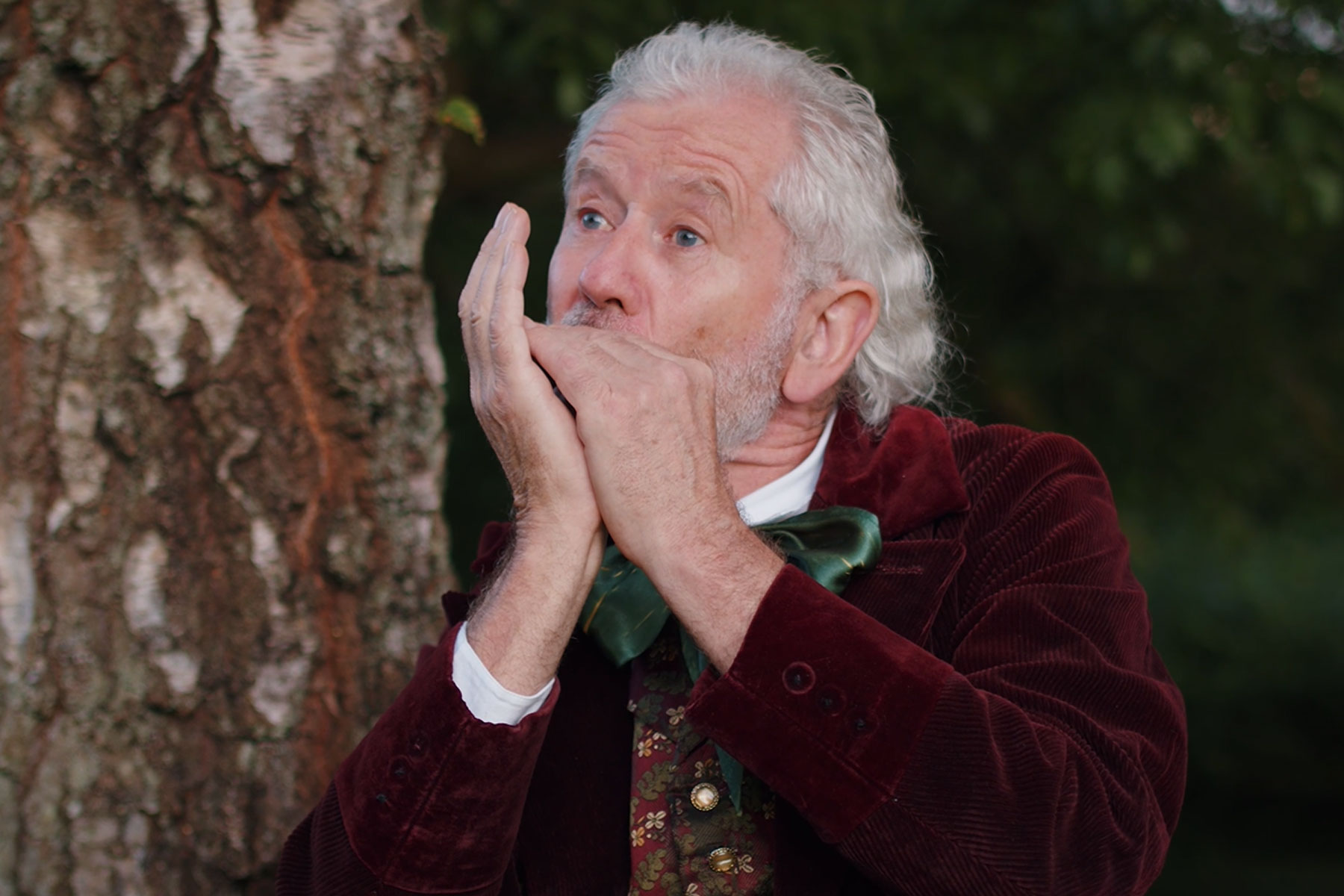 A smartly dressed hobbit plays the harmonica next to a tree