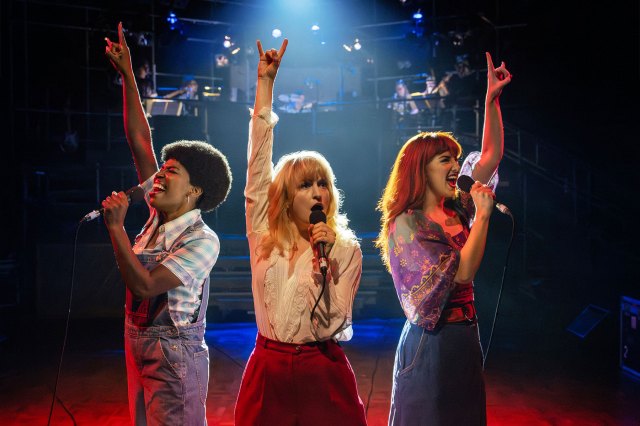 Three female singers stand with microphones in one hand and the other hand raised in the air