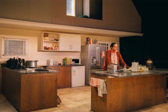 An actress stands in the middle of an onstage kitchen set