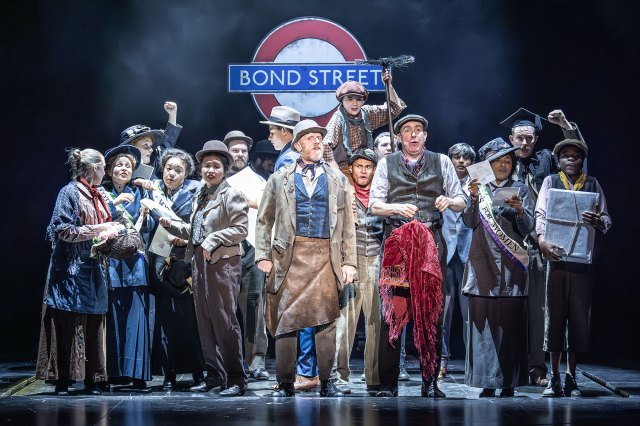 A group of actors in working-class period costumes in front of a Bond Street underground sign