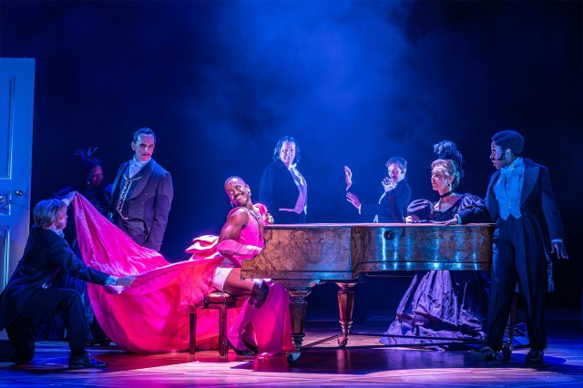 An actor in a pink gown at the piano, surrounded by actors in tuxedos