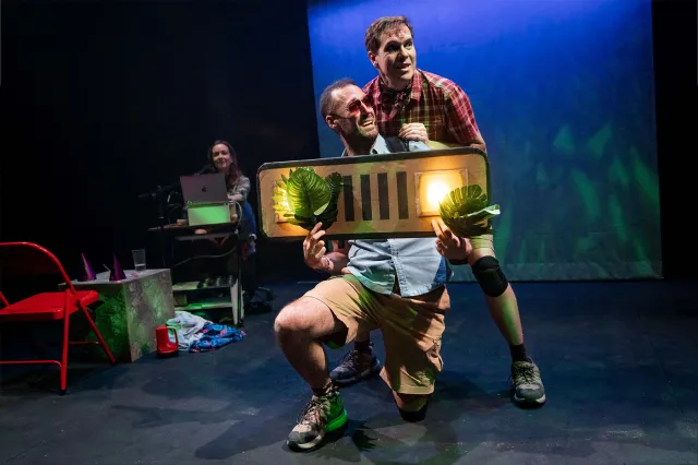 Two actors on stage, one holding the front piece of a jeep with headlights on