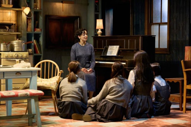 A group of actors on stage, portraying a mother and four daughters sat around a piano