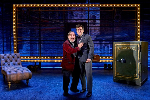 Two actors joyously cling together on stage, with a chaise longue on the left and a large safe on the right
