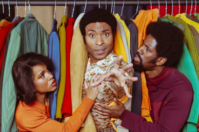 Three actors appearing through a clothes rack of brightly coloured jackets