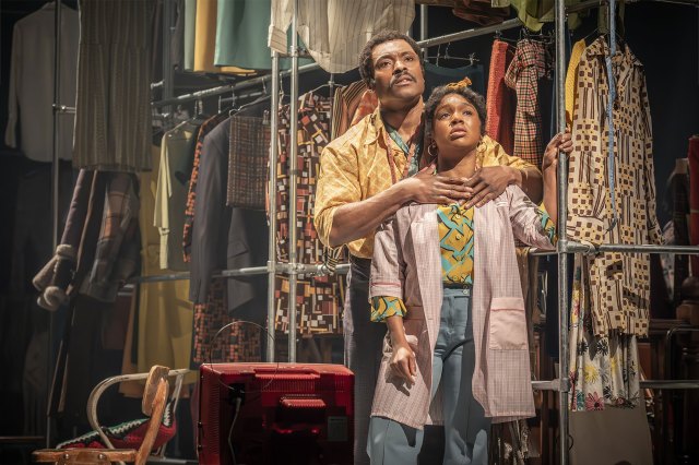 An actor and an actress in Sixties attire embrace in front of racks of clothing