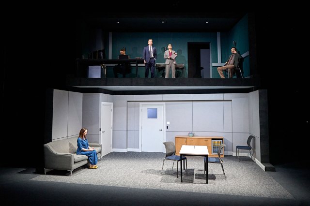 A group of actors on a theatre set that resembles a two-storey office design