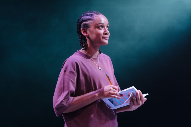 An actress with braided hair on stage, holding a notepad and pen