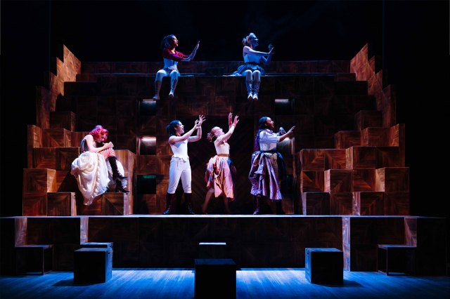 A group of actresses standing on a multi-platform stage in period costumes