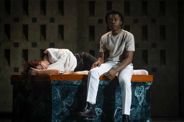 An actor sits on a stone alter next to an actress who lies motionless on stage