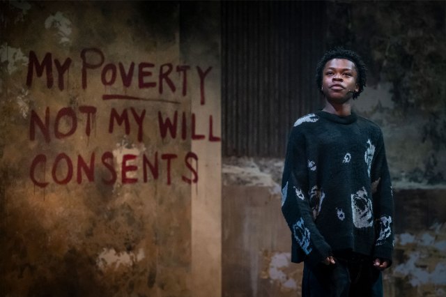 An actor stands on stage by a wall with the words "My Poverty Not My Will Consents" written on it with red paint