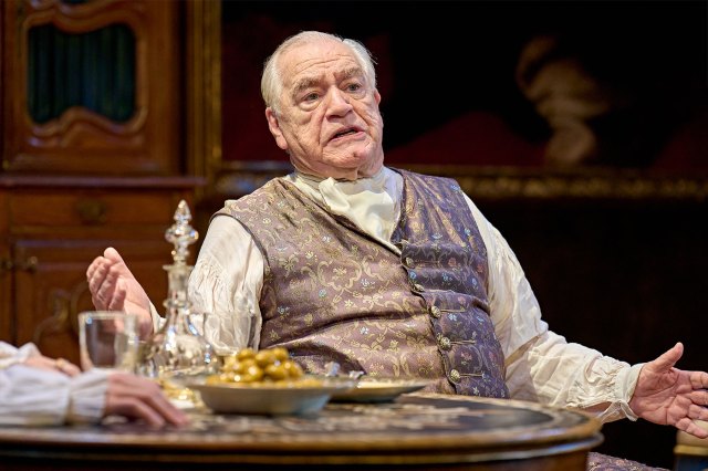 An elderly actor on stage in period costume, sat at a table