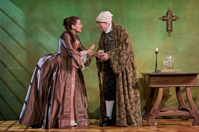 An actress and an actor on stage in period costumes, next to a wooden table and candle with a crucifix hanging on a green wall backdrop