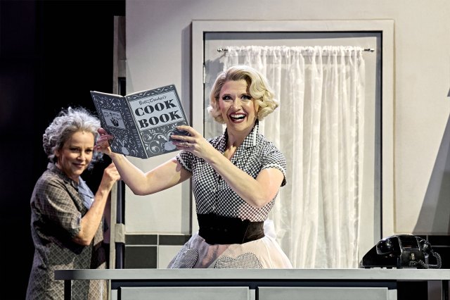 An actress on stage in a 1950's monochrome housewife costume, holding up a cookbook