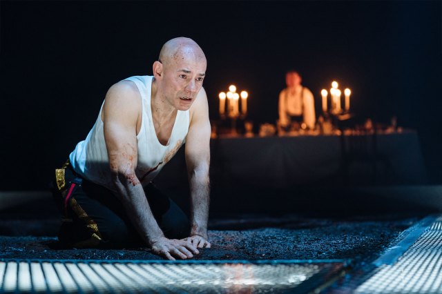 An actor on stage on his hands and knees with a candle-lit altar in the background