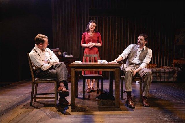 Two actors in 1940s period costumes sit at a table with a woman in a red dress standing behind it and between them