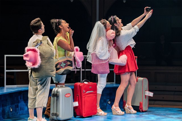 A group of actors on stage, one dressed as a bride-to-be, taking a selfie with travel suitcases at their feet