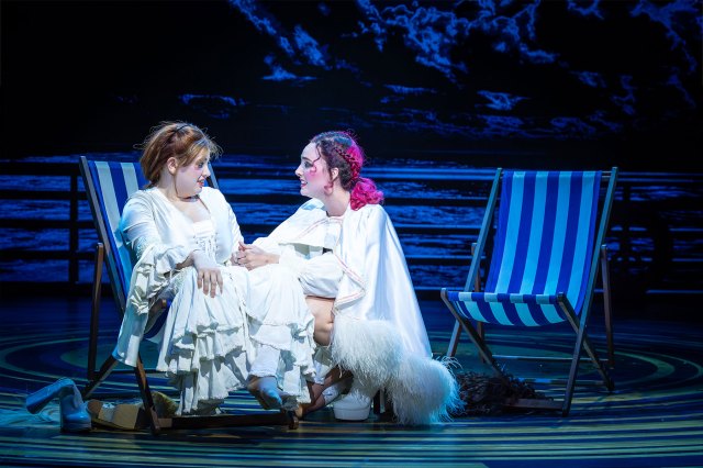 Two actresses on stage, one sat in a deckchair, the other crouching down next to her.