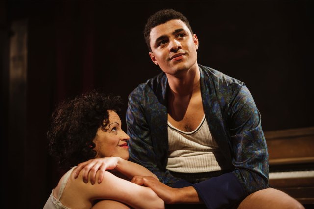 An actress leans her arms on the leg of a seated actor onstage