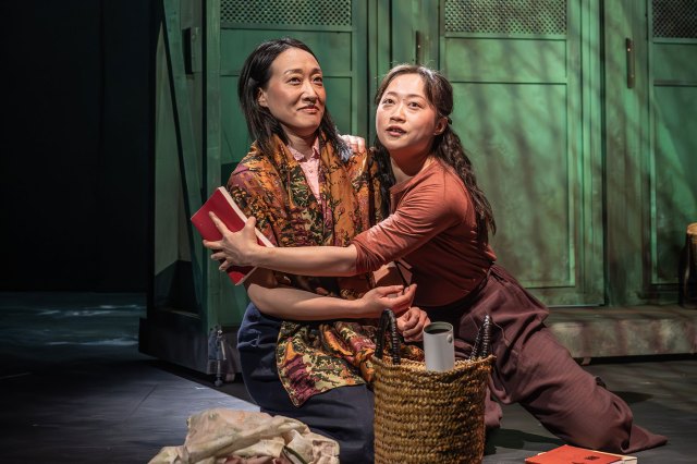 Two actresses are knelt on the stage floor, embracing each other