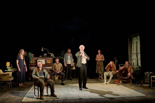 A group of actors gathered on stage on a dimly lit living room set