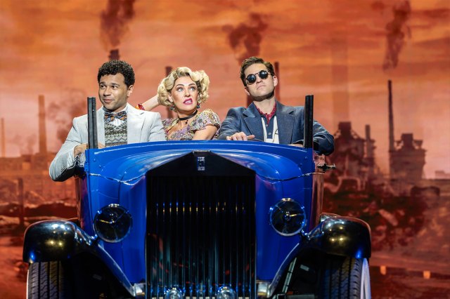 Two actors and an actress in 1920's attire sitting inside a blue 1920's car on stage