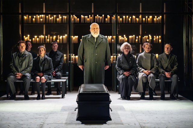 A cast of actors gather on stage with a black coffin in the foreground and rows of lit candles in the background.