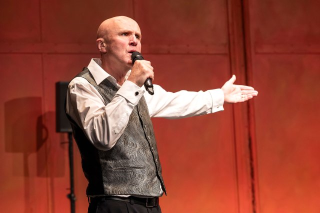 An actor on stage, speaking into a microphone in front of a reddish backdrop, wearing a white shirt, grey waistcoat and black trousers.
