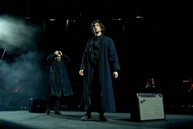 Two actors in black overcoats, standing on stage.