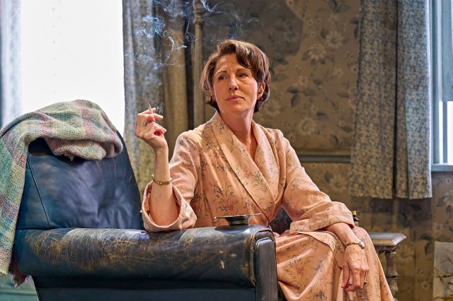 An actress in a dressing gown, sitting on stage in an armchair with a cigarette in her hand.