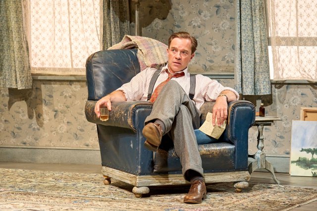 An actor, sitting in an armchair on stage.