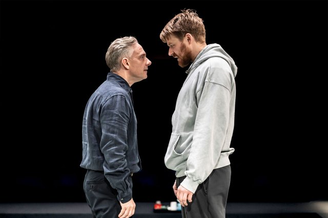 Two actors stand on stage facing each other.