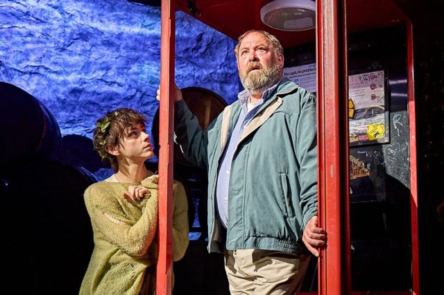 An actor stands on stage inside a red telephone box with a younger actor leaning against it.