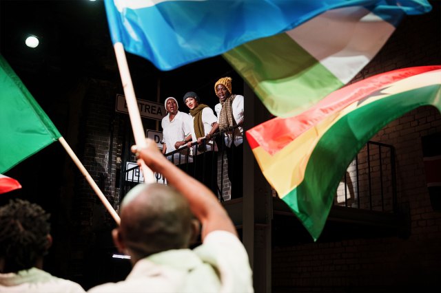 Three actors on a balcony looking down at a crowd of actors waving various flags from African nations.