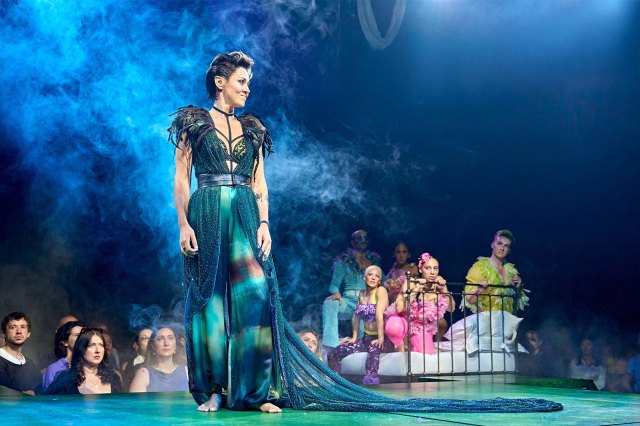 An actress in a majestic green gown on stage with a group of actors sitting on a bed in the background.