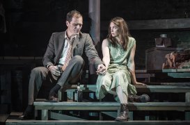 An actor and an actress sit on wooden steps on stage, holding hands.