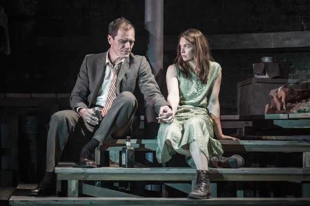 An actor and an actress sit on wooden steps on stage, holding hands.