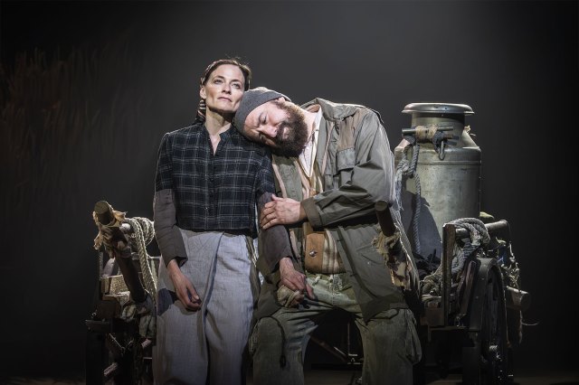 An actor leans his head on an actress' shoulder, both sitting on a wooden milk cart.