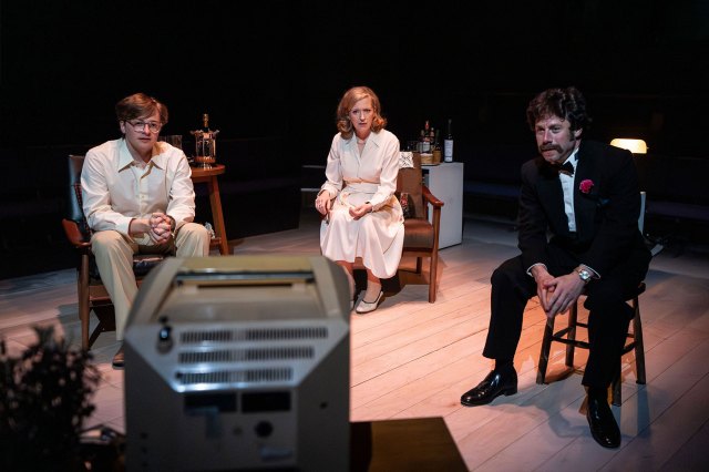 An actress with an actor on each side of her, all sat on stage watching a television set.