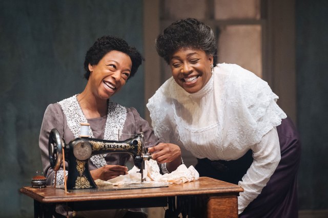 Two actresses in period costumes, gathered around a sewing machine