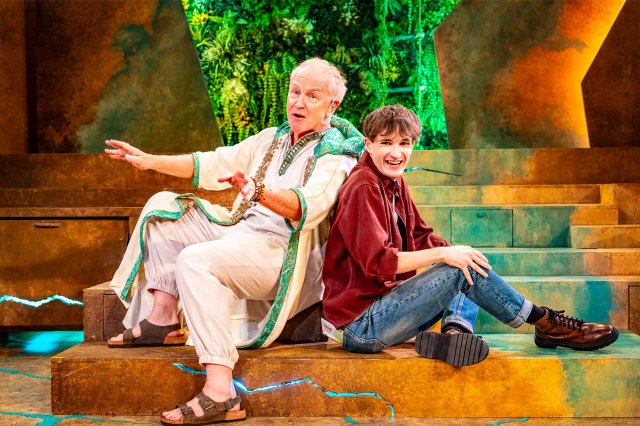 An older actor in thobes and sandals and a young actor in modern attire sit back-to-back on a stage with sandy-coloured steps.