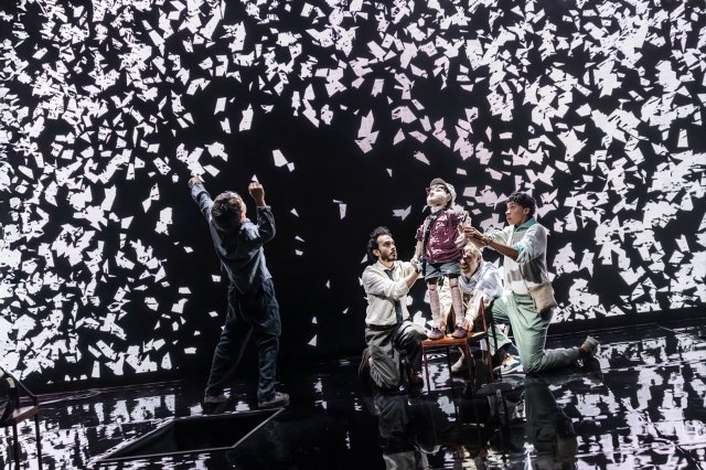 A group of actors on stage with a wooden puppet boy and a video backdrop showing hundreds of flying white birds.